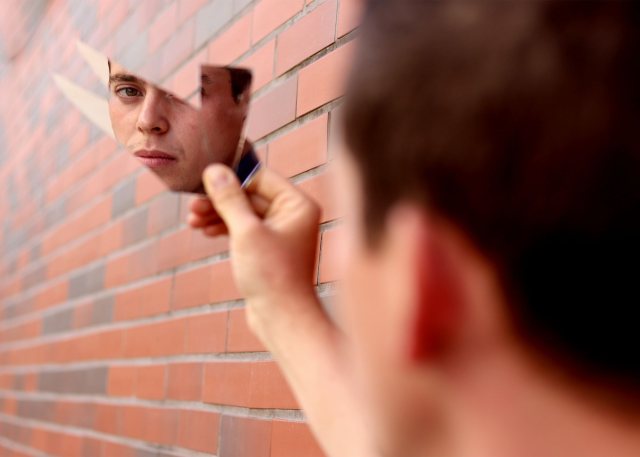 A man's face is reflected in a fragment of a mirror held against a brick wall.